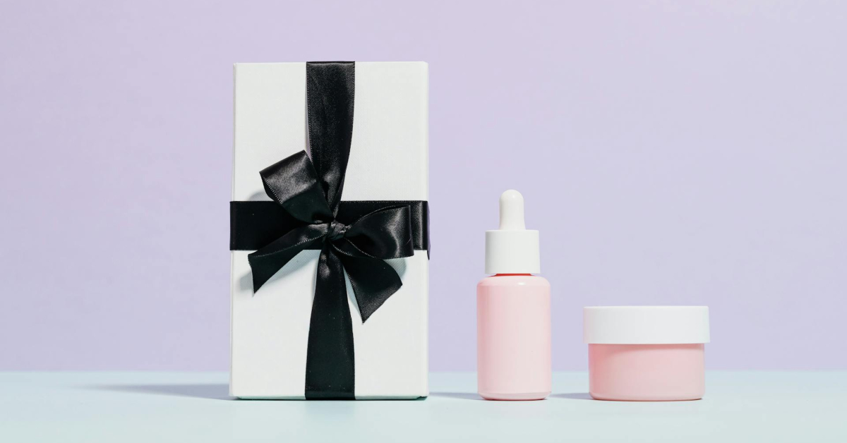 A white gift box with a black ribbon showcases elegant packaging next to a pink dropper bottle and a small pink cosmetic jar on a light blue surface against a pastel purple background.