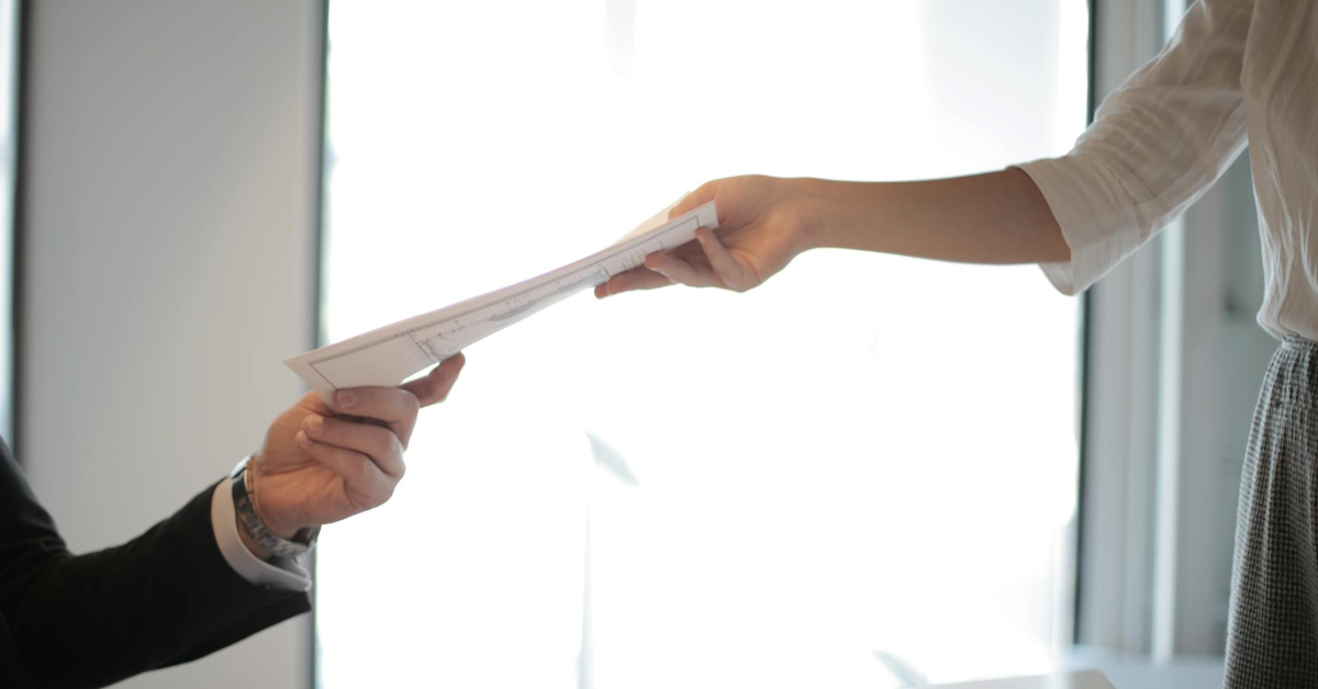 In a brightly lit room, two people connect over a stack of papers, one person handing them over like the key to successful job placements and the other receiving eagerly.