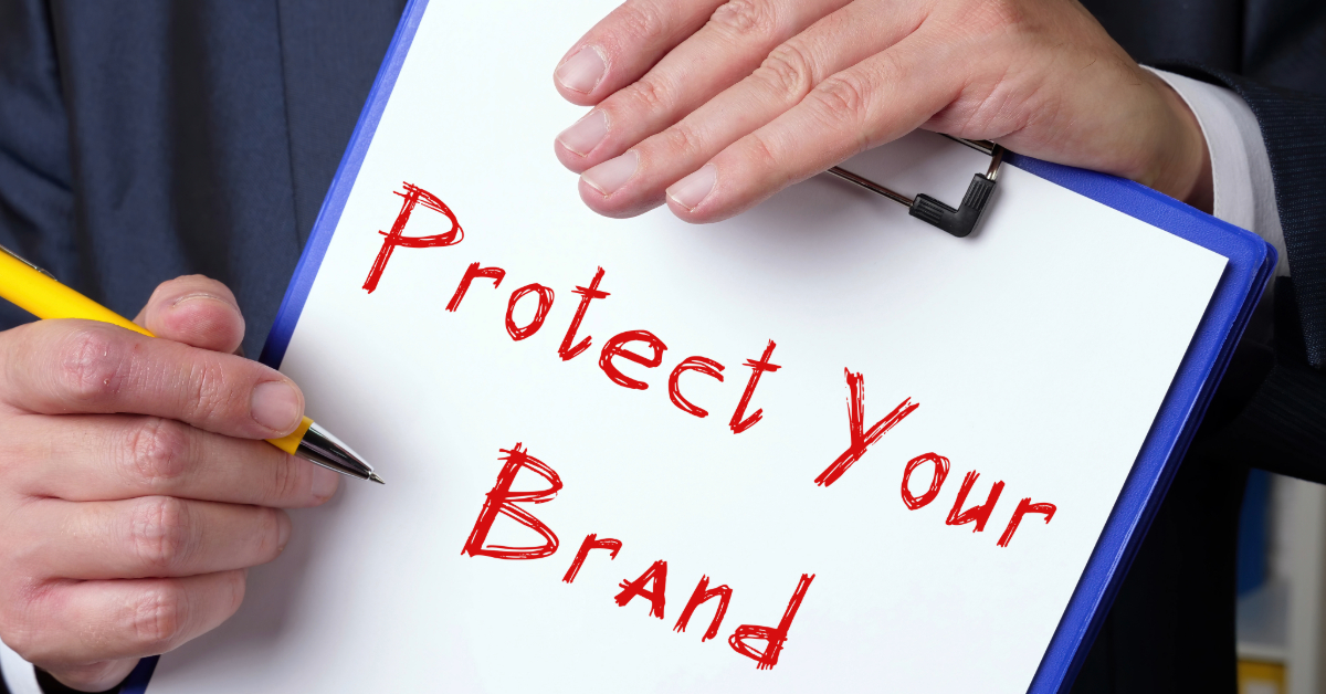 A person holds a clipboard with a sheet of paper that says "Protect Your Brand" in red letters and writes with a yellow pen, emphasizing the importance of brand protection against domain infringement.