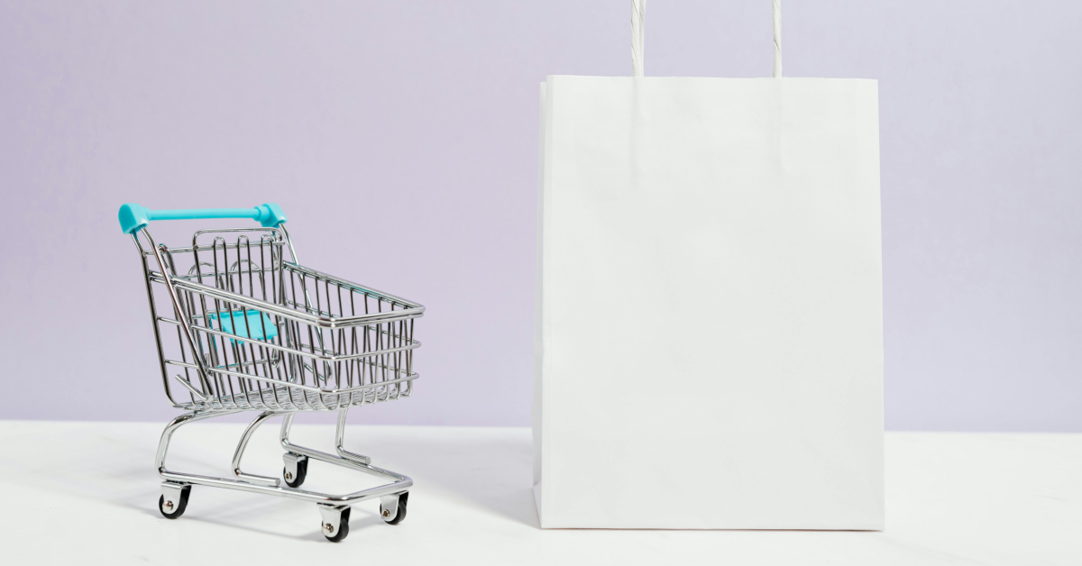 A miniature shopping cart with blue handles is positioned next to a plain white shopping bag, symbolizing online business essentials on a light surface with a pale background.
