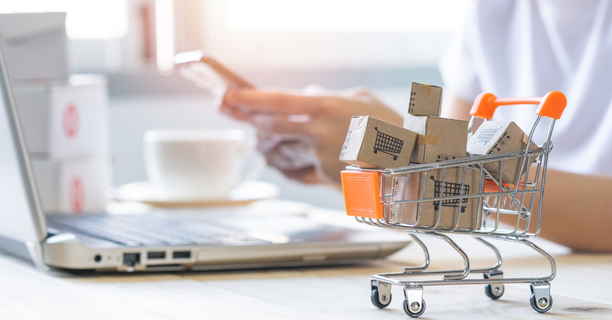 A person using a smartphone and laptop sits at a desk, highlighting the rise of investment opportunities in e-commerce marketplaces, with a mini shopping cart filled with small cardboard boxes in the foreground.