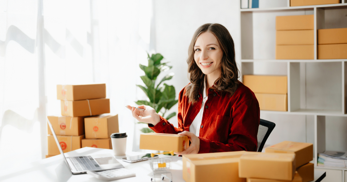 A woman sits at a desk with a laptop, surrounded by cardboard boxes, holding a package and pen—likely preparing shipments for her e-commerce business as she works to build reputation through top strategies.