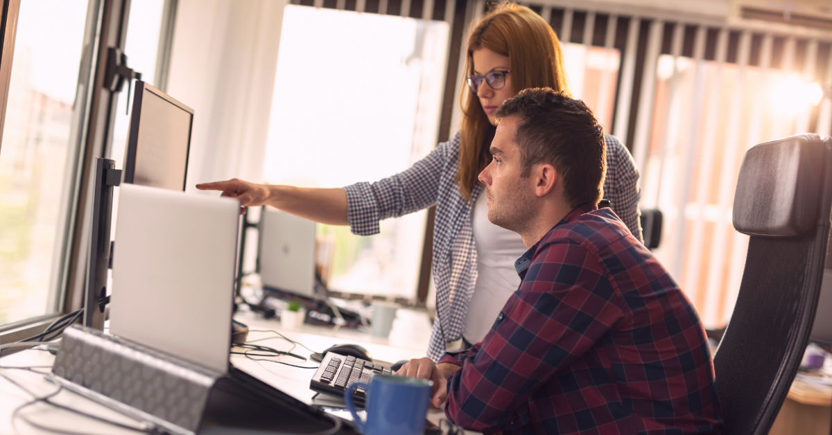 Two people working at a desk in an office; one is sitting at a computer while the other stands beside, pointing at the screen—discussing IT outsourcing options to avoid common IT support mistakes.