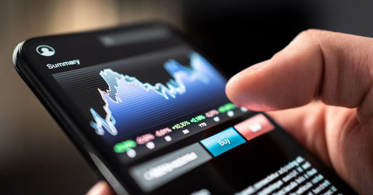 A person uses a smartphone to view and interact with a stock market trading app displaying a fluctuating price chart, buy/sell options, and cryptocurrency trading features for ecommerce investors.