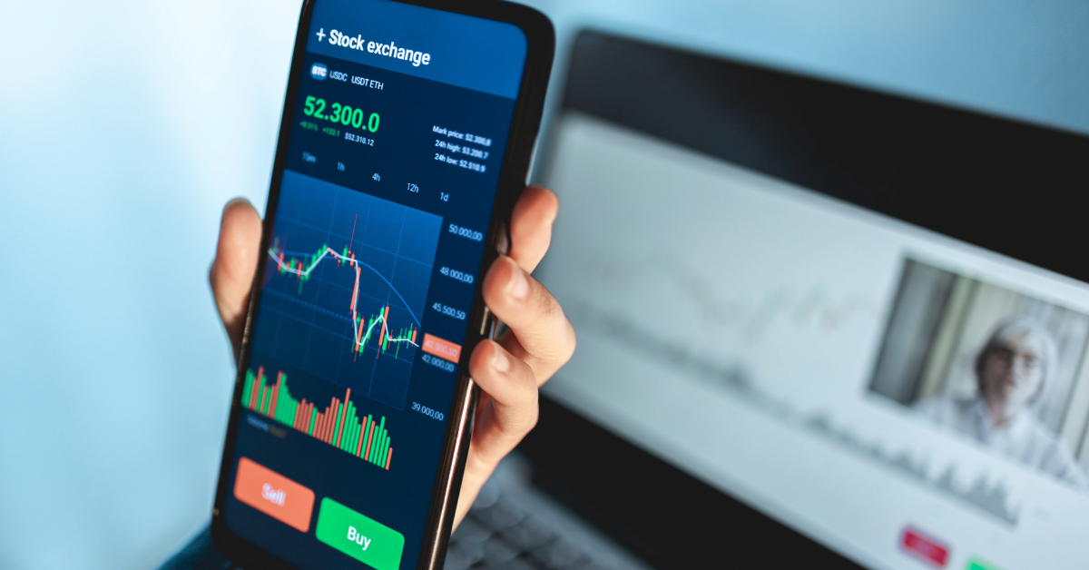 A hand holds a smartphone displaying a stock exchange app with price charts and buy/sell buttons, in front of a laptop showing a blurred screen—capturing the start of an exciting trading journey.