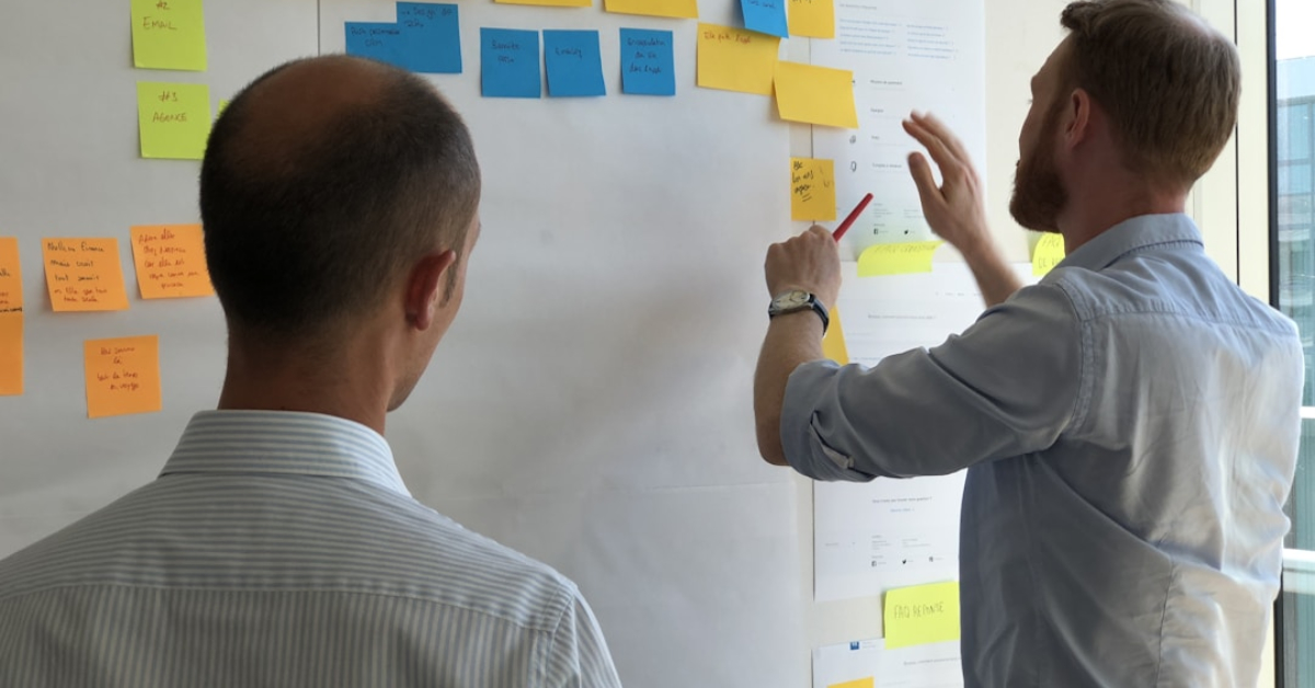 Two men stand in front of a whiteboard covered with sticky notes, discussing the customer journey and arranging notes to highlight the benefits during a collaborative planning session.