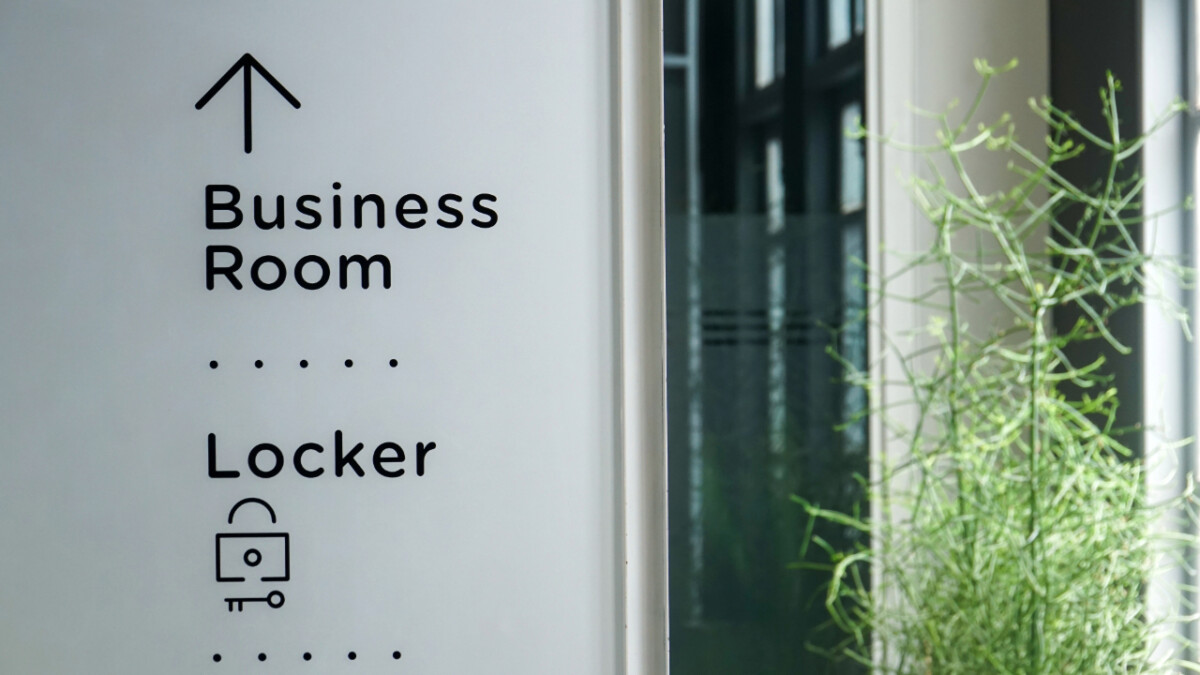 A sign with arrows directs to a business room and locker area; clear office signage enhances navigation, while a plant and windows in the background add to the comfortable office environment.
