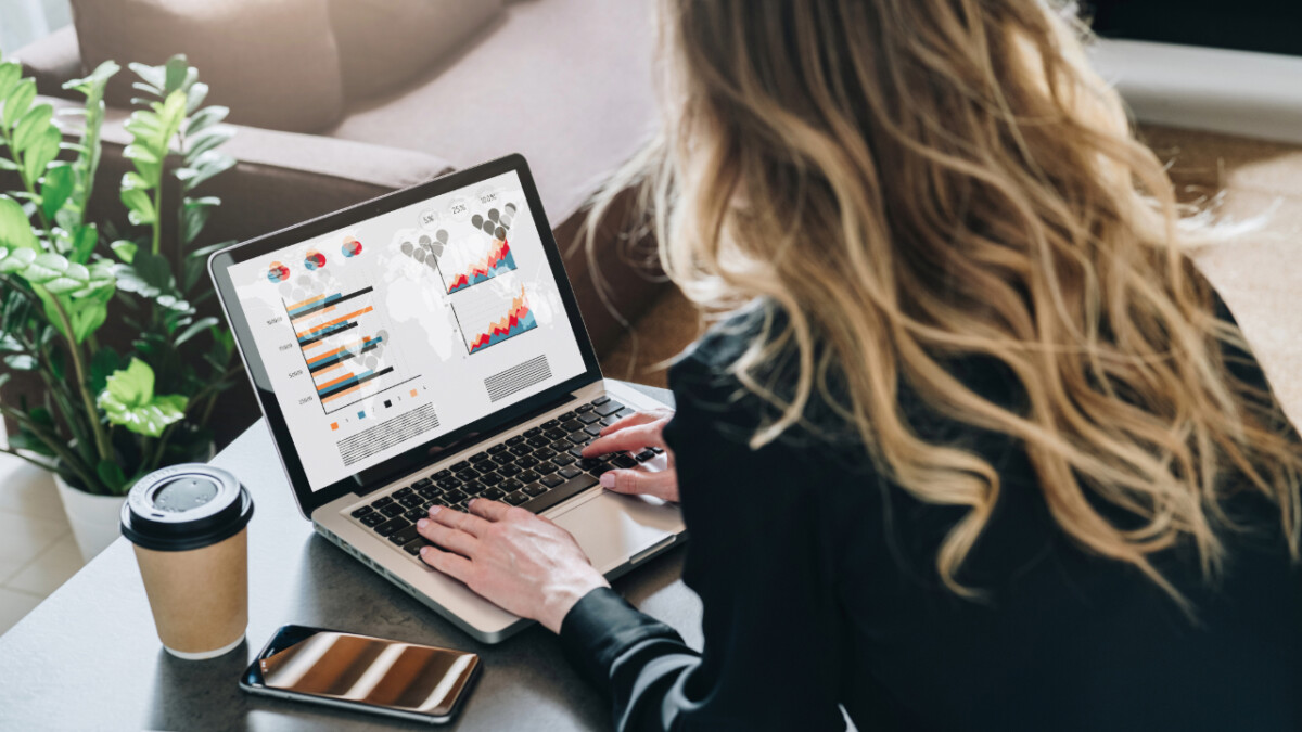 A woman researches What is SEO in ecommerce marketing on her laptop displaying graphs and charts, while a smartphone, coffee cup, and potted plants sit on the table nearby.