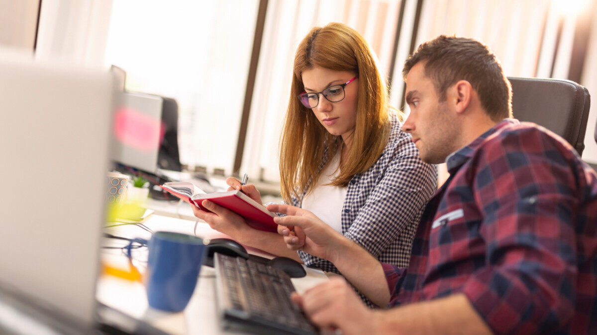 Two people sitting at a desk in an office, looking at a notebook together while working on computers and discussing the benefits of Ecommerce outsourcing.