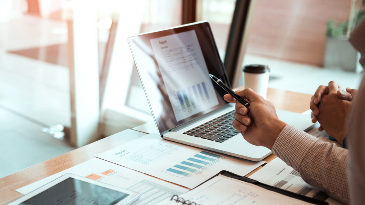 Two people review financial charts and graphs on a laptop and printed sheets at a desk in a well-lit office, discussing financial strategies for their ecommerce business.