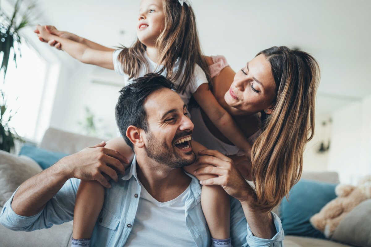 A man carrying a smiling young girl on his shoulders, with a woman beside them, all appearing happy and playful in a bright living room, reflecting the peace of mind that comes with the best 2024 term insurance.