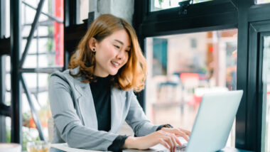 A woman in a gray blazer is sitting at a table, typing on her laptop and smiling, clearly enjoying her work as an SEO expert, with large windows in the background.
