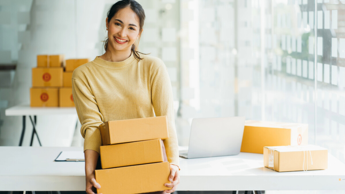 A woman stands in an office holding a stack of cardboard boxes—likely shipping out Top Selling Products on Shopify in 2024—while a laptop and more boxes sit on the table behind her.
