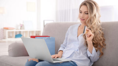 A woman sits on a couch using a laptop, holding a credit card with shopping bags beside her, embodying the latest 2024 eCommerce trends in online retail.