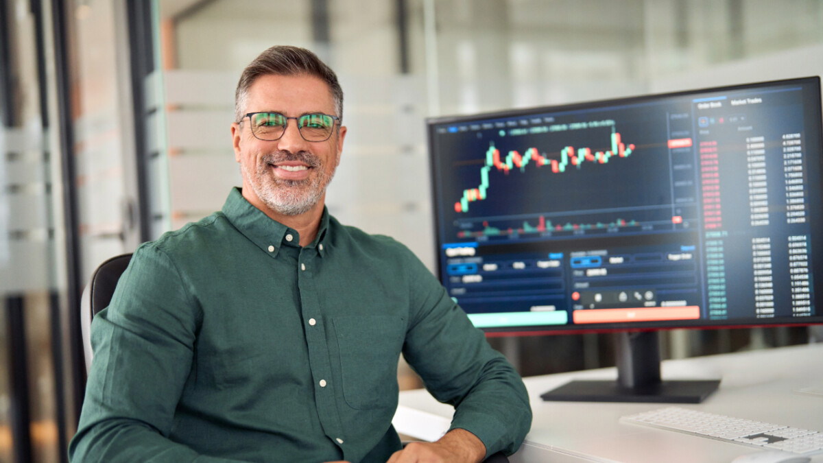 A man sits at a desk smiling, with a computer monitor beside him displaying financial trading charts and data from his preferred cryptocurrency spot brokerage.