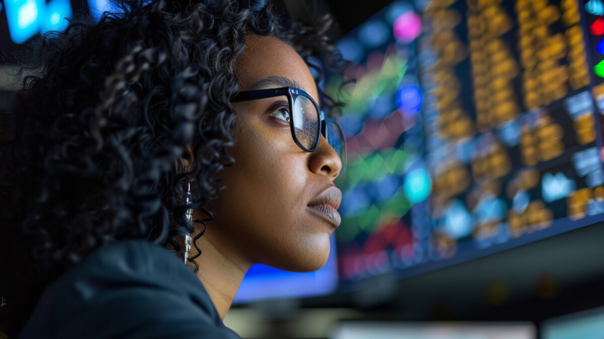 A woman wearing glasses looks intently at multiple screens displaying colorful financial data and stock charts in a control room, analyzing investing opportunities for Canadians.