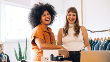 Two women standing together in a clothing store, smiling at the camera, with boxes and clothes racks visible in the background—perfect inspiration for anyone looking to start a dropshipping business.