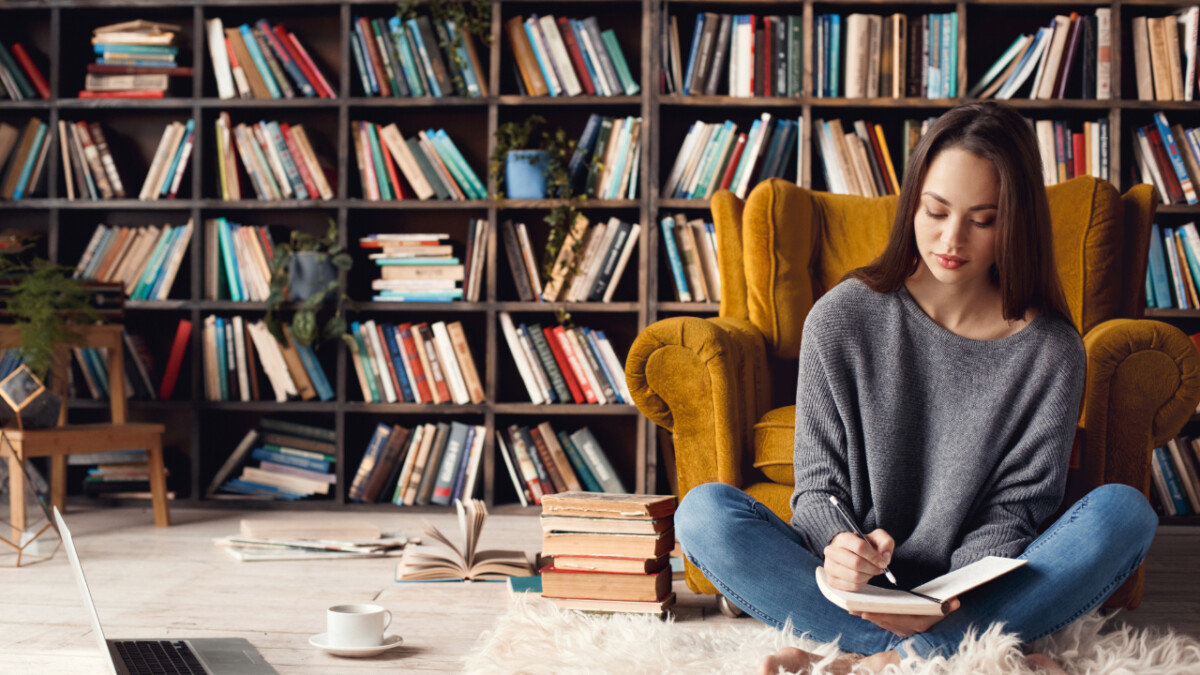 A woman sits cross-legged on a rug, writing in a notebook—perhaps inspired by creative writing careers—with a yellow armchair, bookshelves filled with books, and a cup on the floor nearby.