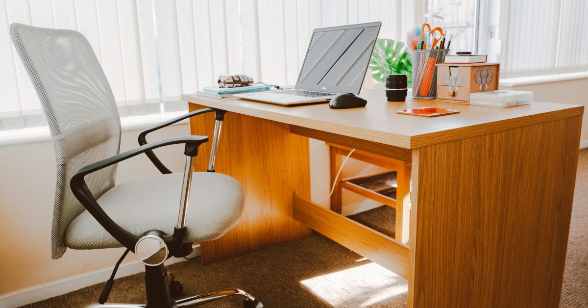 A modern wooden desk with a laptop, office supplies, a potted plant, and an ergonomic chair in a bright room with vertical blinds—perfect for focusing on financial success or launching other ventures.