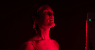 A person stands with eyes closed under a Red Light Therapy Device, facing a vertical LED panel in a dark room.