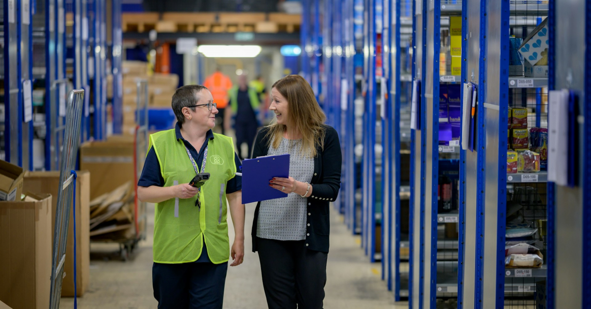 Two workers walk and talk in a warehouse aisle, discussing warehouse management. One wears a safety vest and holds a scanner; the other carries a clipboard. Shelves with various products line both sides of the aisle.