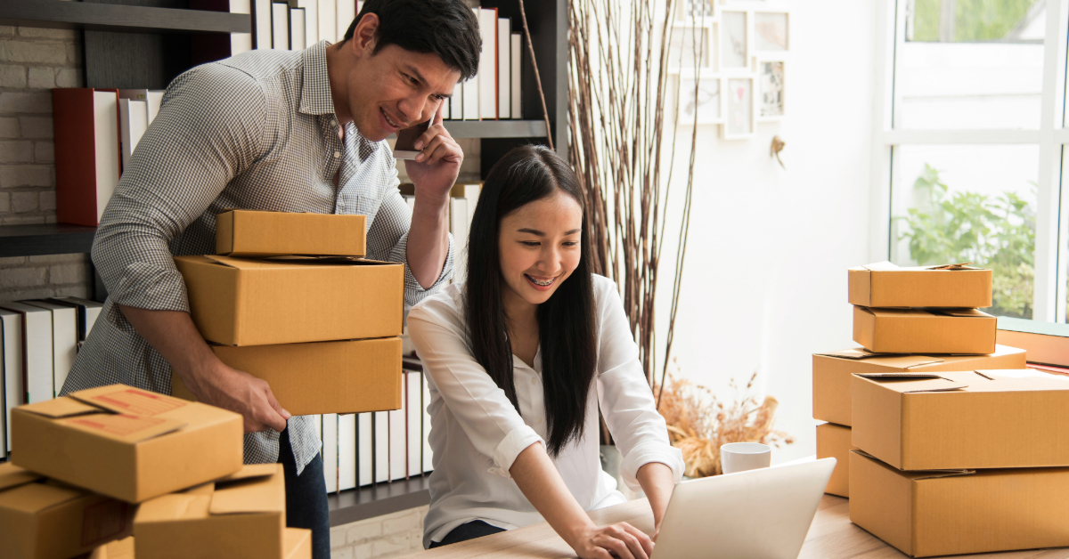 Two people work together in a room filled with cardboard boxes, running their online business; one is on the phone and holding boxes while the other uses a laptop to manage different types of online business tasks.