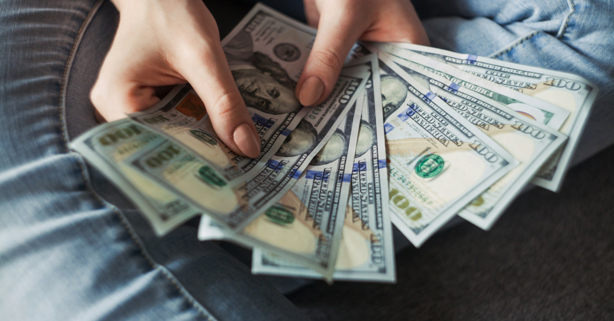 A person is holding and fanning out several $100 U.S. dollar bills while sitting on the floor, showcasing the rewards of discovering ways to make money online.