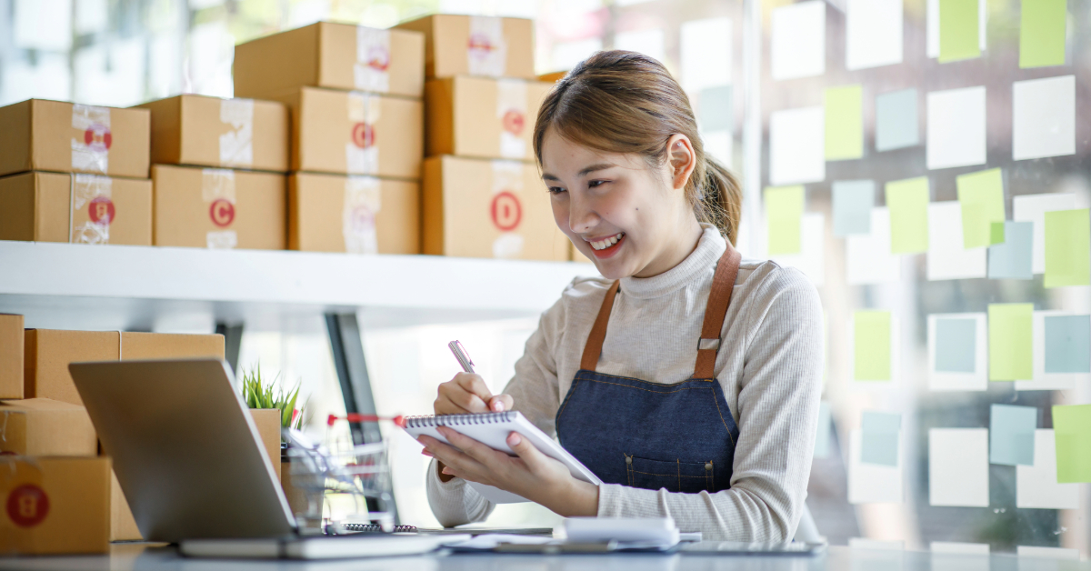 A woman in an apron sits at a desk with a laptop, writing in a notebook, surrounded by cardboard boxes and sticky notes as she reviews a retail pricing guide.
