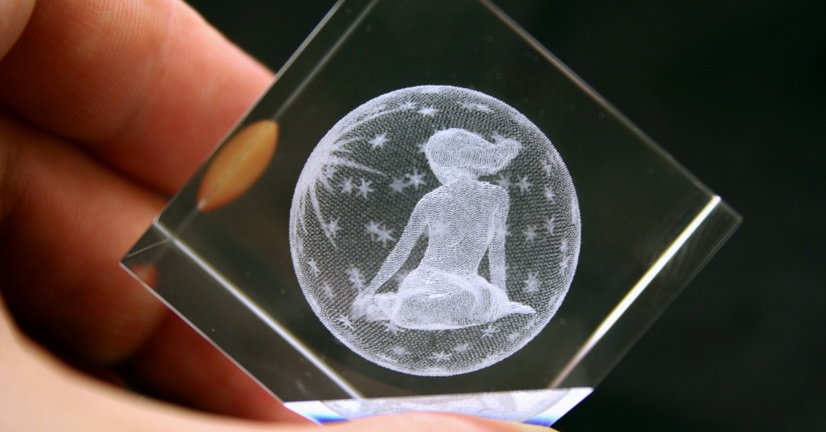 A hand holds a clear glass cube showcasing a 3D crystal engraving of a seated person surrounded by stars inside a circular border.