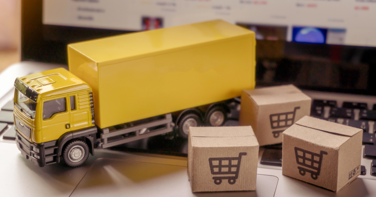 A yellow toy truck and small cardboard boxes with shopping cart icons are placed on a laptop keyboard, highlighting e-commerce and delivery concepts, as well as the importance of choosing the right logistics carrier.
