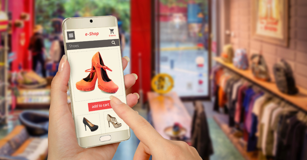 A person explores footwear options on a mobile app while standing in a clothing store, showcasing the benefits of ecommerce with shelves of clothes and a window visible in the background.