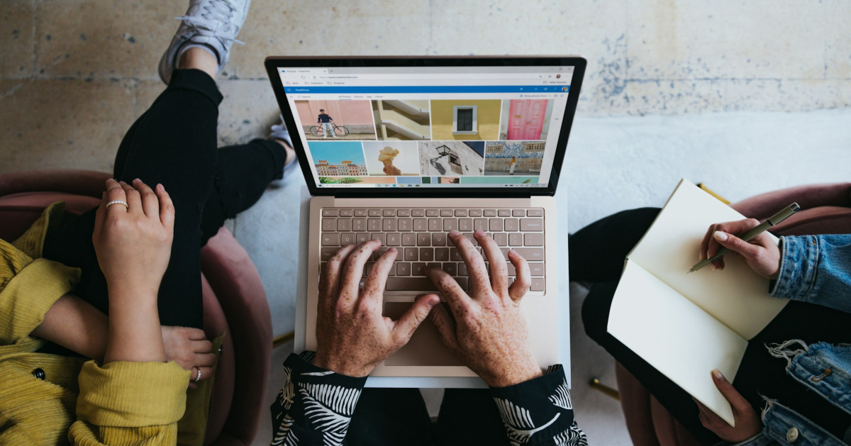 Three people sit together; one person types on a laptop displaying photo thumbnails, while another writes in a notebook, discussing strategies to increase visibility for a new company.
