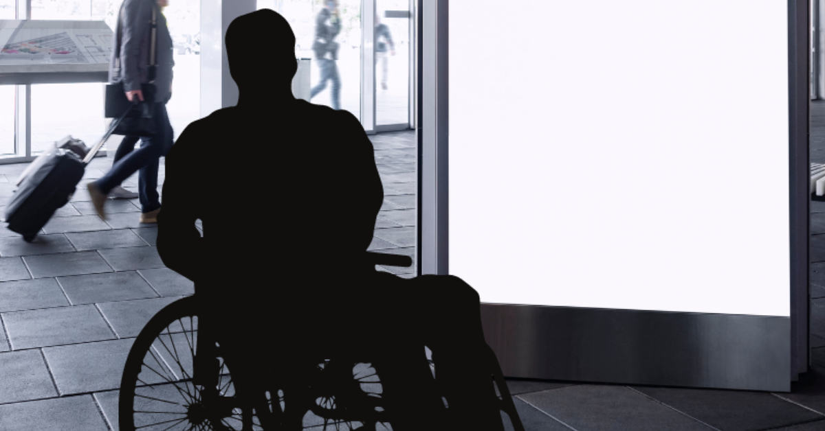 A person in a wheelchair is silhouetted against a large, bright accessible digital signage panel in an indoor public space while people walk by in the background.