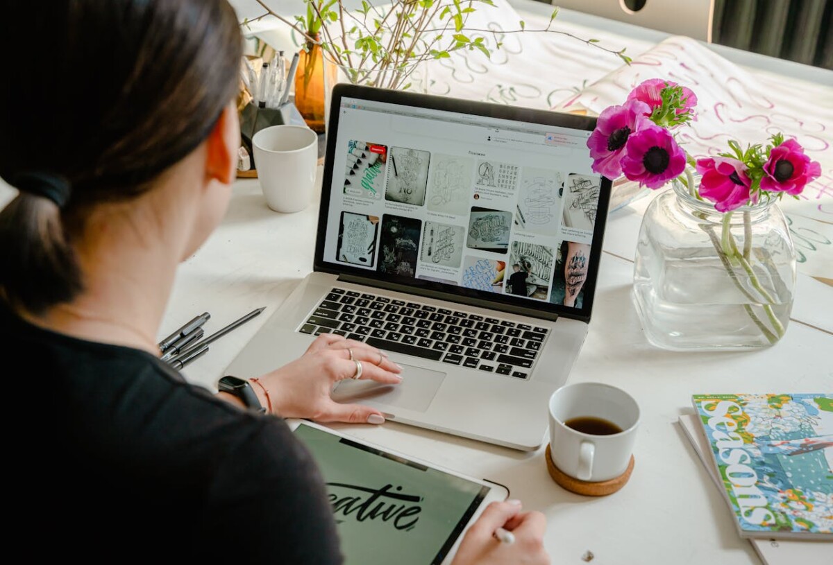 A person working at a desk with a laptop displaying design images, an iPad, coffee, a vase with flowers, and a magazine—preparing to break down technical concepts for better audience engagement.