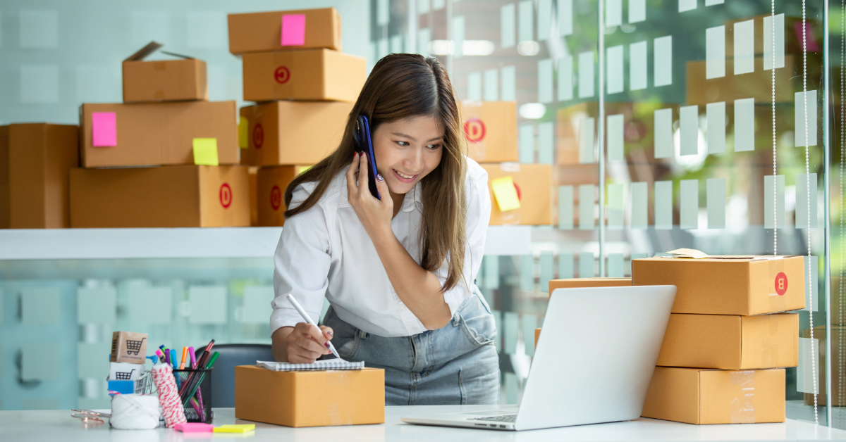 A woman speaks on the phone while writing notes and looking at a laptop, surrounded by cardboard boxes in a modern office—a scene typical for scaling eCommerce stores in 2024.