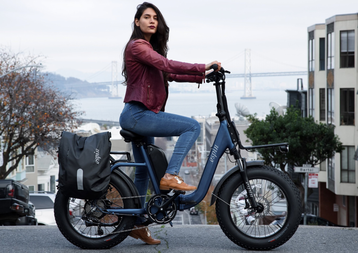 A woman in a maroon jacket and jeans sits on an electric bicycle, used for last mile delivery, with a bag attached against a cityscape and bridge backdrop.