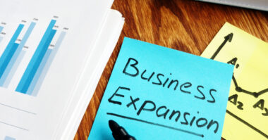 A sticky note labeled "Business Expansion" beside financial graphs and a pen on a wooden desk, perfect for those looking to expand your business through effective business preparation.