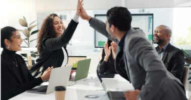 Two business professionals high-five during a meeting, celebrating a Classic Gem direct marketing success, while others at the table smile and applaud; laptops and documents are visible on the table.