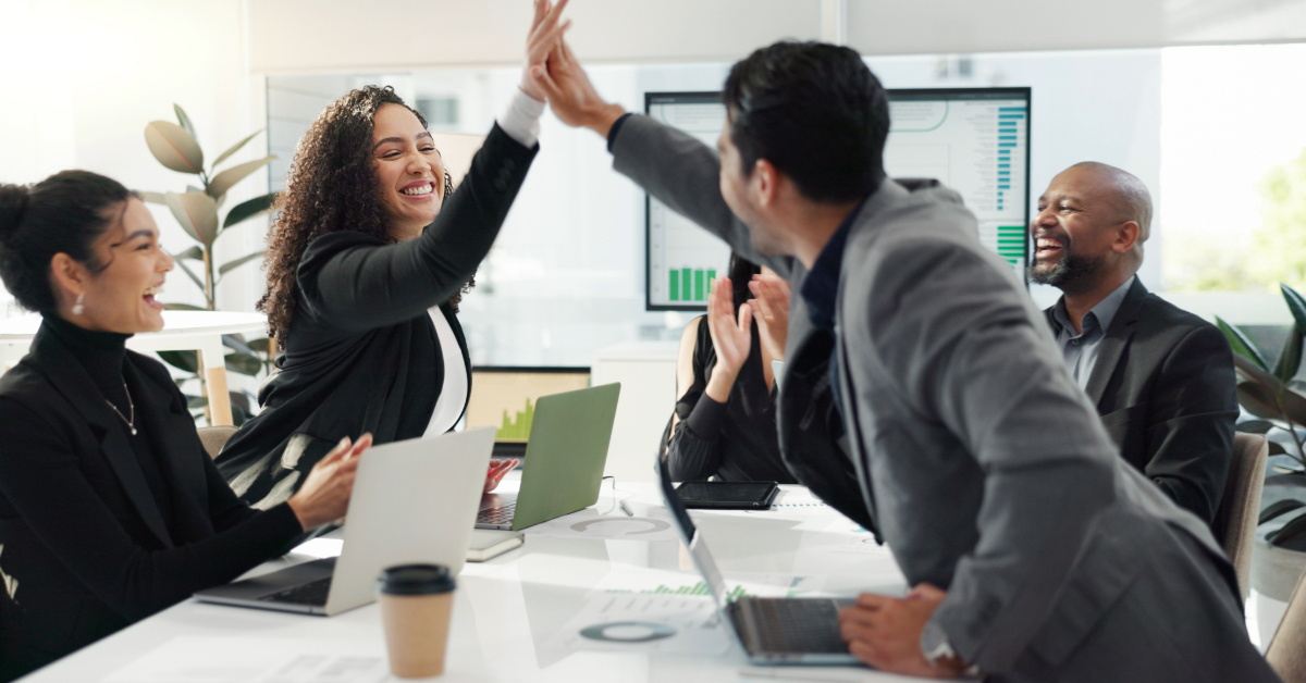 Four business professionals sit at a conference table, two exchanging a high five while others smile, surrounded by laptops and documents as they discuss brand consistency and marketing strategies.