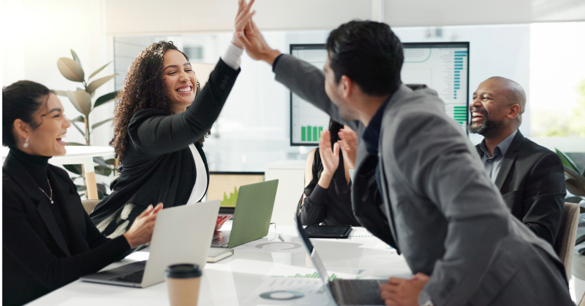 Two business professionals high-five during a meeting, celebrating a Classic Gem direct marketing success, while others at the table smile and applaud; laptops and documents are visible on the table.