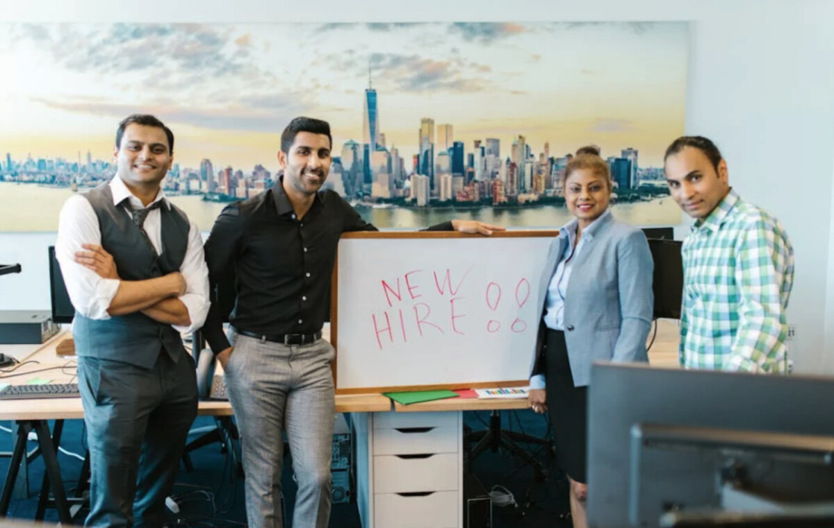 Four professionally dressed people stand in an office next to a whiteboard with "NEW HIRE!!!" written on it; a cityscape mural is in the background, showcasing the excitement to hire developers and build dynamic teams in 2024.