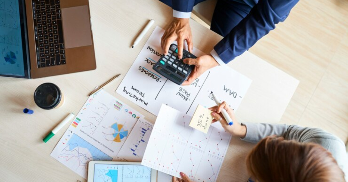 Two people review charts and graphs at a desk, using a calculator, tablet, and laptop; papers with data and a coffee cup are also visible—perfect for anyone planning to start an e-commerce business or seeking a complete guide 2024.