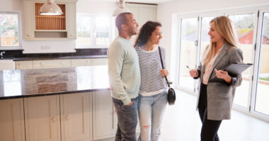 A couple stands in a modern kitchen, smiling and talking with a real estate agent holding a clipboard, discussing the latest house buying trend.