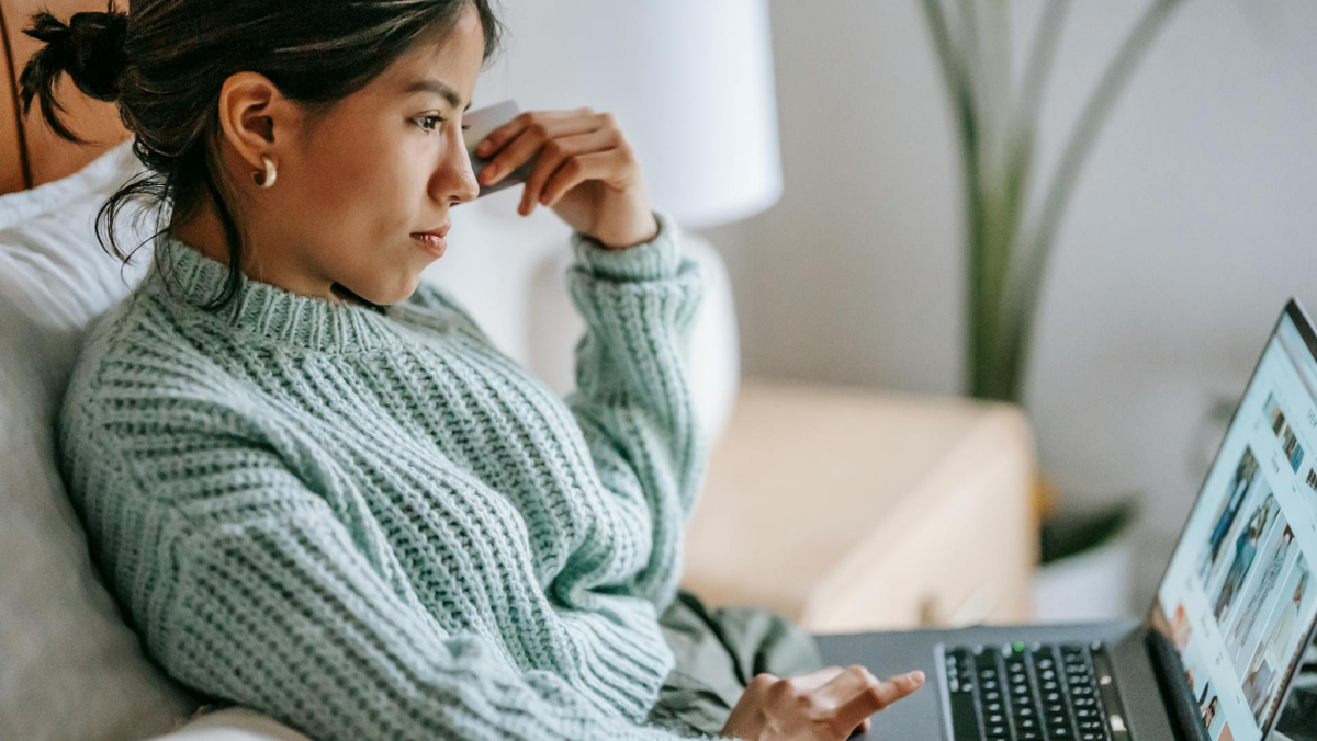 A person in a green sweater sits on a bed, browsing a website on their laptop and exploring how the modern world benefits consumers.