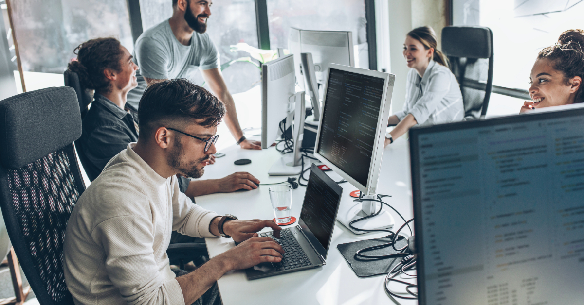 Five people in an office setting work at desks with computers—some typing, some talking—as large windows provide natural light, reflecting a team focused on streamlining operations with tailored technology solutions.