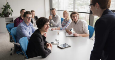 A group of people sit around a conference table in a modern office, listening to a woman at the head of the table as she demonstrates technical leadership and charts the course for their global journey.