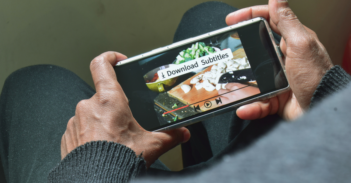 Person holding a smartphone and watching a cooking video with an on-screen prompt reading “Download Subtitles.” Chopped vegetables are visible on a cutting board, highlighting the convenience of automatic captioning for recipe transcription.