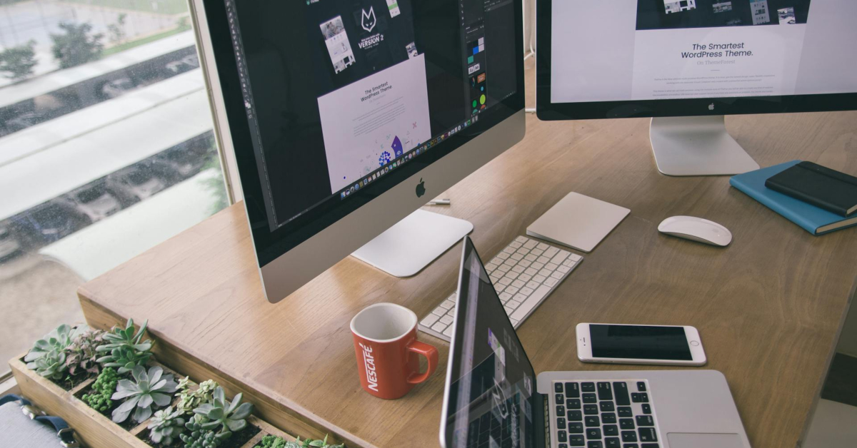 A modern workspace designed for business growth, featuring two desktop monitors, a laptop, smartphone, notebook, keyboard, mouse, coffee mug, and a small indoor succulent garden on a wooden desk.