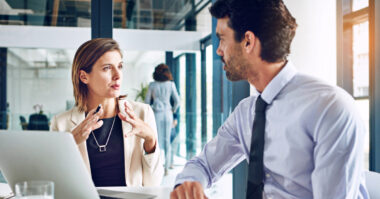 Two business professionals discuss Odoo benefits at a desk with a laptop in a modern office, exploring how Odoo for Retail can streamline their operations.