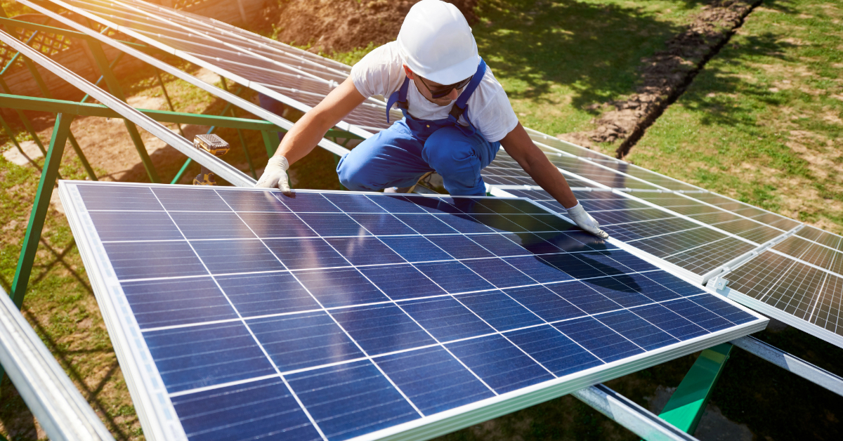 A worker in a white helmet and gloves installs a solar panel outdoors on a metal frame under sunlight, contributing to solar energy and powering the future.